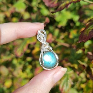 May include: A wire-wrapped pendant featuring a round, iridescent blue-green labradorite gemstone encased in silver-coloured metal wire. The wire forms a decorative loop at the top for suspension, with intricate coils securing the stone in place.