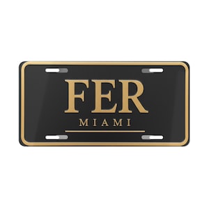 Puede incluir: Placa de matrícula negra con borde y letras doradas. La placa dice "FER" encima de "MIAMI" en una fuente en negrita. La placa tiene cuatro ranuras para el montaje.
