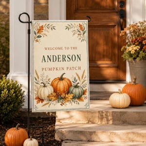Puede incluir: Una bandera de jardín rectangular colgada en un soporte de metal negro. La bandera presenta un fondo crema con ilustraciones otoñales de calabazas, trigo y hojas. Muestra el texto: WELCOME TO THE ANDERSON PUMPKIN PATCH.