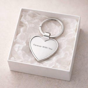 Puede incluir: Un llavero plateado en forma de corazón con la inscripción "Forever With You" se presenta en una caja de regalo blanca. El llavero tiene un anillo grande y está sobre papel de seda blanco.