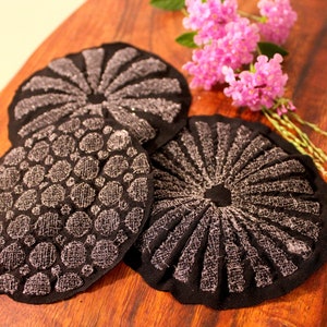 May include: Three black fabric coasters with silver embroidery. The coasters feature different designs, including a circle of dots, a sunburst pattern, and a circle of radiating lines.