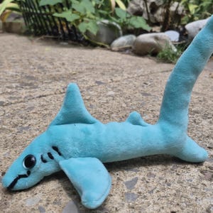 May include: A light blue plush shark toy with a soft, fuzzy texture, featuring large black plastic eyes, embroidered black gill slits on the sides, and a long, upright tail fin. The stuffed animal is positioned horizontally on a textured surface.