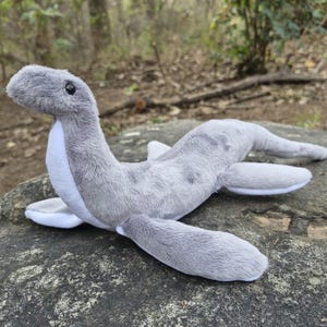 Op de afbeelding: Een pluche speelgoed van een grijs zeemonster, mogelijk het Monster van Loch Ness, met een lange nek, kleine zwarte ogen en vinnen. Het speelgoed heeft een witte buik en ligt op een rots.