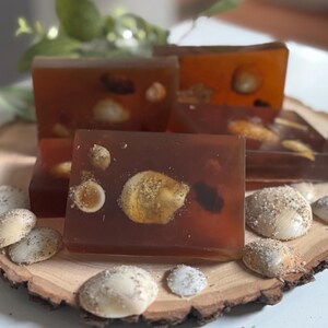 May include: Four amber-colored soap bars with embedded seashells, arranged on a wooden slice. The soaps are surrounded by small seashells. The overall theme is a beach or coastal aesthetic.