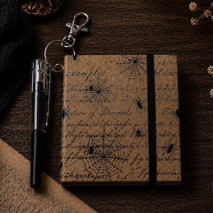 May include: A square notebook with a tan cover featuring black spiderweb and spider illustrations, cursive text, and a black elastic band. A black pen is attached to the notebook via a silver metal keychain ring and a lobster clasp.