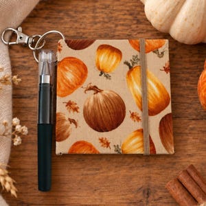 May include: A square notebook with a beige cover featuring a repeating pattern of orange and brown pumpkins and small leaves. The notebook includes a black pen attached to a silver metal keychain ring and a vertical elastic band closure.