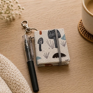 May include: A small, rectangular notebook with a cream-colored fabric cover featuring a pattern of dark gray, blue, and orange mushrooms. The notebook is held closed by a gray elastic band and is attached to a silver metal keychain with a black pen.