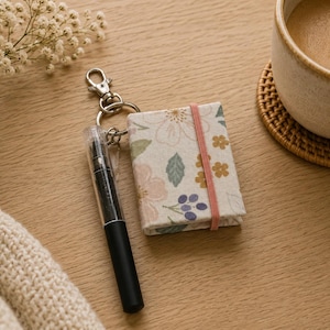 May include: A miniature rectangular notebook with a cream-colored cover featuring a floral pattern of pink, yellow, and blue flowers and green leaves. A pink elastic band secures the notebook, which is attached to a silver metal keychain with a black pen.