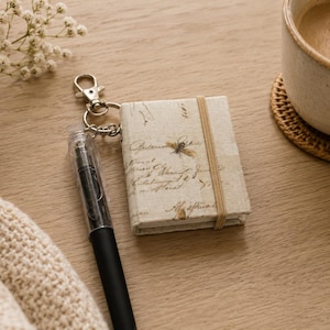 Pode incluir: Um pequeno caderno em estilo vintage com uma ilustração de abelha e texto manuscrito, preso a um porta-chaves. Uma caneta preta e uma chávena de café também estão na superfície de madeira. O caderno é mantido fechado por uma faixa elástica bege.
