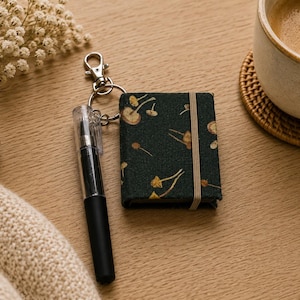 May include: A small dark green notebook with a mushroom pattern, secured by a vertical elastic band, attached to a silver metal keyring with a black and clear plastic pen. The notebook cover features a repeating print of small golden-brown mushrooms.