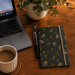 May include: A dark green notebook featuring a repeating pattern of small brown and tan mushrooms, secured with a vertical elastic band. The notebook is positioned next to a black pen with a metal clip and a silver keychain attachment.