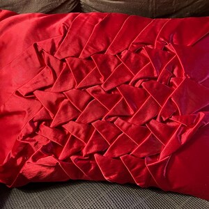 May include: A red velvet pillow with a unique braided design. The pillow is soft and plush, and the braided design adds a touch of texture and dimension.