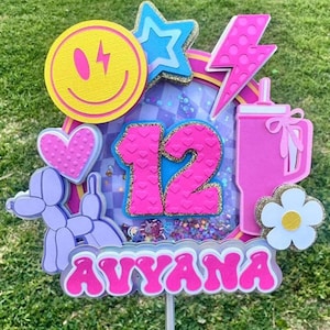 May include: A pink and purple cake topper featuring the number 12, the name Avyana, and a variety of colourful decorations, including a smiley face, a star, a lightning bolt, a heart, a balloon animal, and a pink cup with a straw.