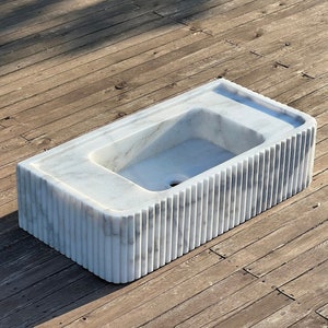May include: A white marble sink with a rectangular shape and a fluted design on the front. The sink is sitting on a wooden surface.