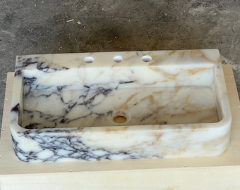 Puede incluir: Un lavabo rectangular de mármol con un patrón veteado crema y marrón. El lavabo tiene tres orificios para grifos y un orificio de desagüe. Está colocado en una caja de madera de color claro.
