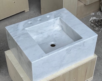May include: A rectangular white marble sink with a built-in basin and three faucet holes. The sink has a polished finish and is set on a wooden crate. The marble has gray veining throughout.