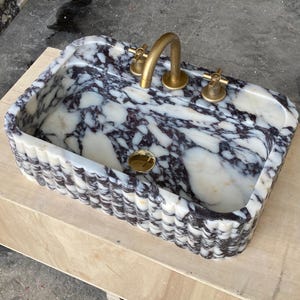 May include: Rectangular marble sink with a white, black, and purple pattern. The sink has a fluted edge and a gold tap and drain. The sink is sitting on a wooden surface.