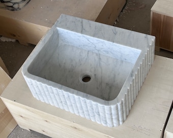 May include: A white marble sink with a fluted edge. The sink is rectangular and has a drain hole in the center. The sink is sitting on a wooden surface.