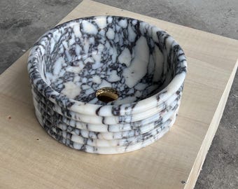 Puede incluir: Un lavabo de mármol blanco con vetas negras. El lavabo es redondo y tiene un diseño en capas. El lavabo está hecho de piedra natural y tiene un acabado liso.