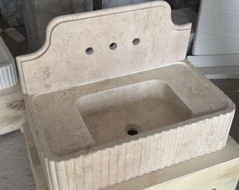 Pode incluir: Uma pia de pedra bege com um protetor de respingos decorativo e uma base canelada. A pia tem três furos para torneiras e um furo de drenagem.