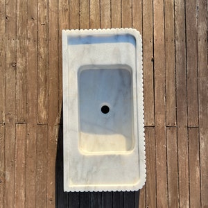 May include: A white marble sink with a scalloped edge. The sink is set against a wooden background.