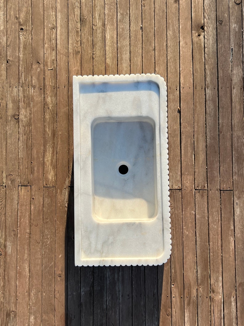 May include: A white marble sink with a scalloped edge. The sink is set against a wooden background.