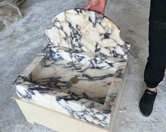 Puede incluir: Un lavabo de mármol blanco con un salpicadero decorativo. El lavabo está montado sobre una base de madera.