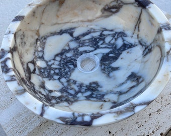 Puede incluir: Un lavabo de mármol blanco con vetas negras y marrones. El lavabo es redondo y tiene un acabado liso y pulido.