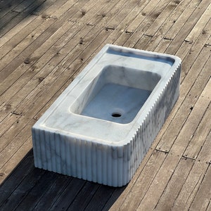 May include: A white marble sink with a rectangular shape and a fluted edge. The sink is set on a wooden surface.