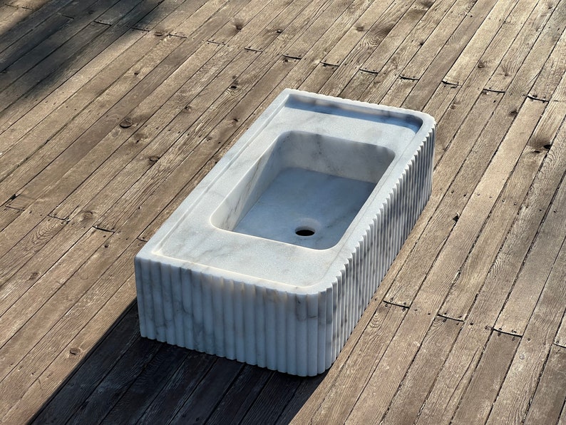 May include: A white marble sink with a rectangular shape and a fluted edge. The sink is set on a wooden surface.