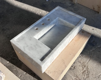 May include: A white marble bathroom sink with three holes for faucets. The sink is rectangular and has a slightly curved edge. It is sitting on a wooden base.