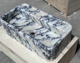 May include: A white marble sink with black veining. The sink has a rectangular shape with a fluted edge. The sink is sitting on a wooden crate.