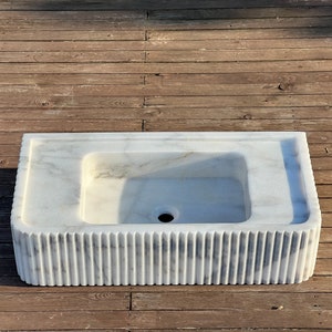 May include: A white marble sink with a rectangular shape and a fluted base. The sink has a single drain hole in the centre.