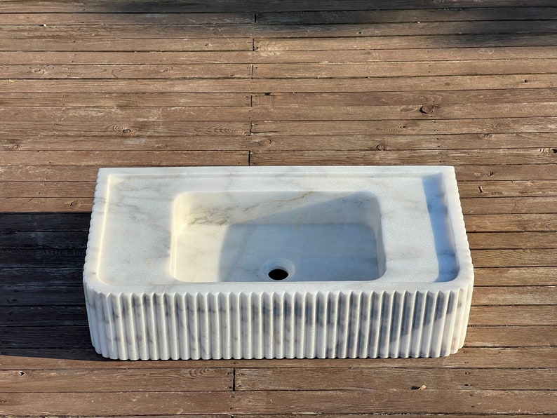 May include: A white marble sink with a rectangular shape and a fluted base. The sink has a single drain hole in the centre.
