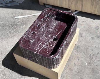 May include: A dark red marble sink with a rippled edge, sitting on a wooden base. The sink has three holes for faucets and a drain.