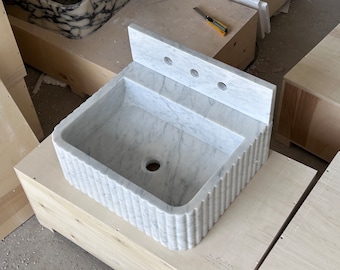 May include: A white marble bathroom sink with a rectangular basin and a fluted exterior design. The sink has a backsplash with three faucet holes. The sink is sitting on a wooden surface.