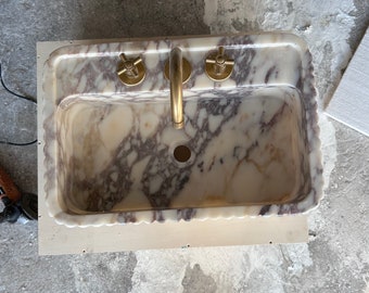 Könnte beinhalten: Ein rechteckiges Waschbecken aus Marmor mit cremefarbenem und violettem Adernmuster sowie einem gewellten Rand. Das Becken verfügt über ein zentrales Abflussloch und eine messingfarbene Armatur mit zwei Kreuzgriffen auf der hinteren Ablage.