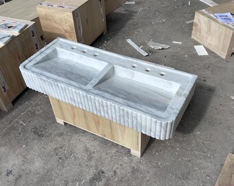 May include: A white marble double sink with a wooden base. The sink has a rippled edge and two separate basins.