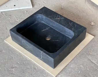Op de afbeelding: Een rechthoekige zwarte marmeren wastafel met witte adering. De wastafel heeft een afgerond interieur en een centraal afvoergat. Het staat op een lichtgekleurde rechthoekige basis en toont een modern badkamerelement.