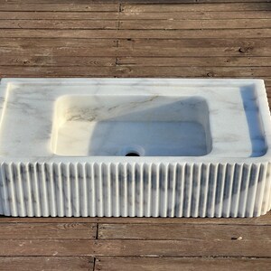May include: A white marble sink with a rectangular basin and a fluted base. The sink is on a wooden surface.