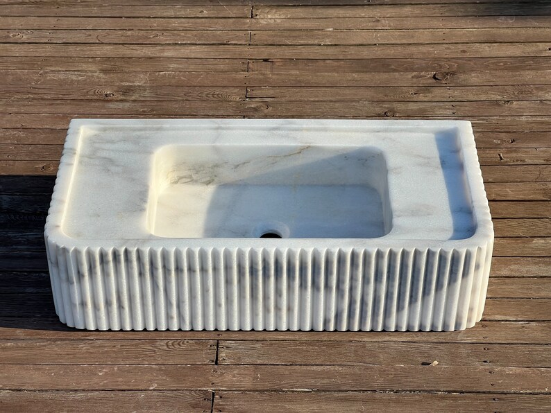 May include: A white marble sink with a rectangular basin and a fluted base. The sink is on a wooden surface.