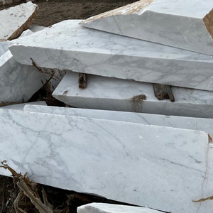 May include: A pile of large, white marble slabs with grey veining. The slabs are stacked on top of each other, and some are partially covered in dirt.