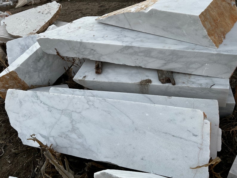 May include: A pile of large, white marble slabs with grey veining. The slabs are stacked on top of each other, and some are partially covered in dirt.