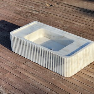 May include: A white marble sink with a rectangular shape and a fluted design on the sides. The sink is sitting on a wooden surface.