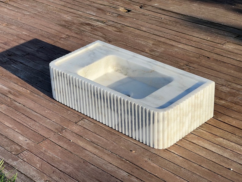 May include: A white marble sink with a rectangular shape and a fluted design on the sides. The sink is sitting on a wooden surface.