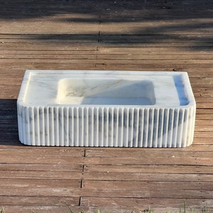 May include: A white marble sink with a rectangular shape and a fluted design around the bottom edge. The sink is sitting on a wooden surface.