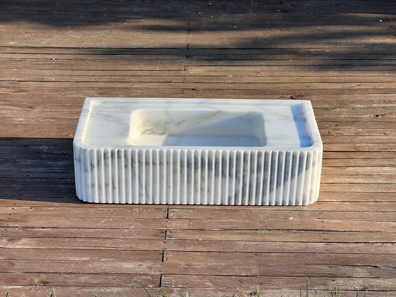 May include: A white marble sink with a rectangular shape and a fluted design around the bottom edge. The sink is sitting on a wooden surface.