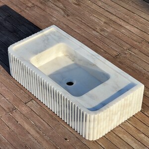 May include: A white marble sink with a rectangular shape and a fluted edge. The sink has a single drain hole in the centre.