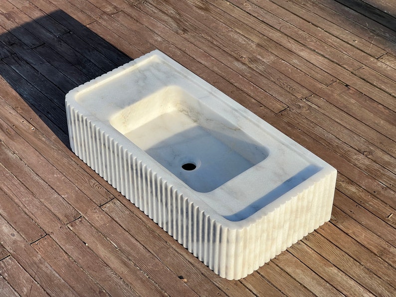 May include: A white marble sink with a rectangular shape and a fluted edge. The sink has a single drain hole in the centre.