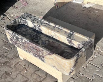 Puede incluir: Un lavabo rectangular de mármol con un patrón de marmoleado marrón claro, rosa y morado. El lavabo está montado sobre una base de madera.